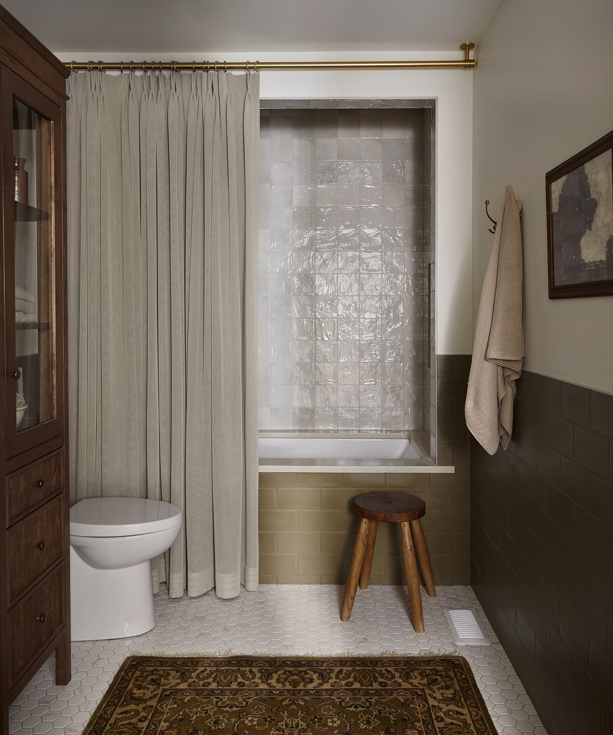 a bathroom with a tall beige shower curtain, zellige tile shower, green tiled walls, a wooden cabinet, and a wooden stool