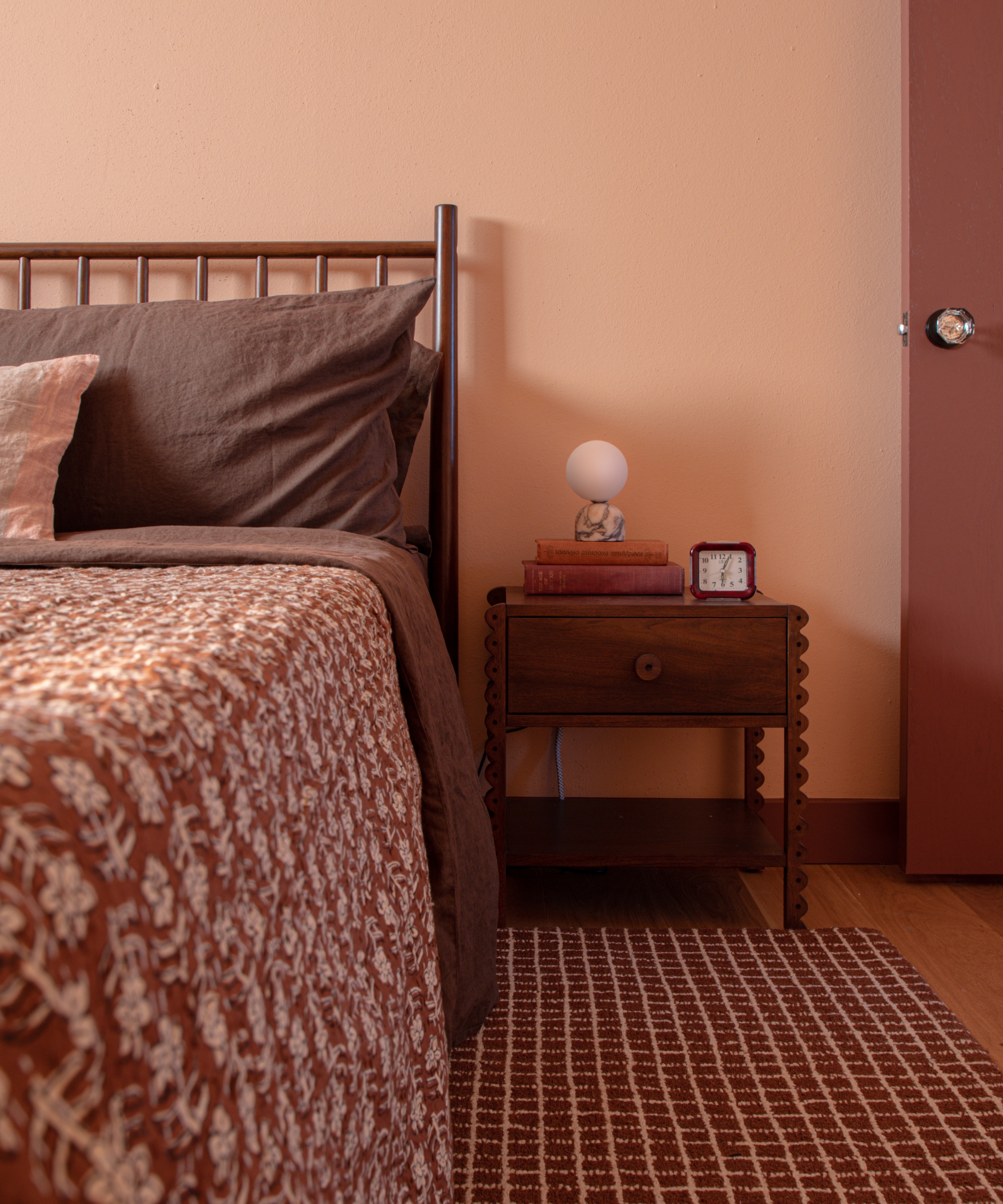 Peach-toned bedroom with brown linen bedding, a patterned rust bedspread, dark wood nightstand, globe lamp and gridded rug.