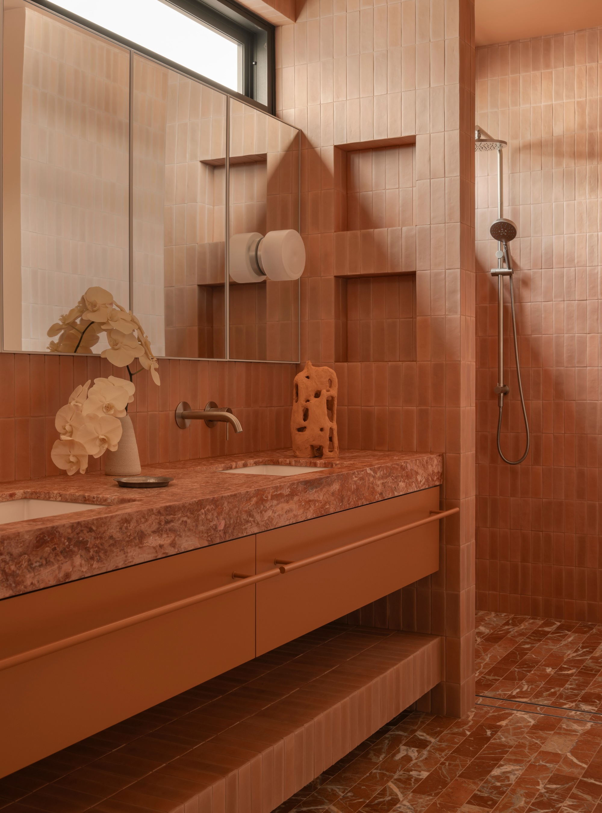 Modern terracotta-toned bathroom with a pink marble vanity, white orchids, vertical stacked wall tiles, large mirror cabinets, and a walk-in shower with reddish marble floor tiles.