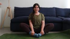 Marina Leiva García sits on a yoga mat performing a butterfly pose. She is sitting in front of a couch, with her knees bent and out to the sides and the soles of her feet touching. She holds her ankles in her hands.