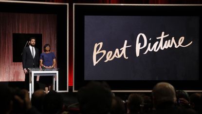 A woman and man stand on stage next to a large screen displaying "Best Picture."