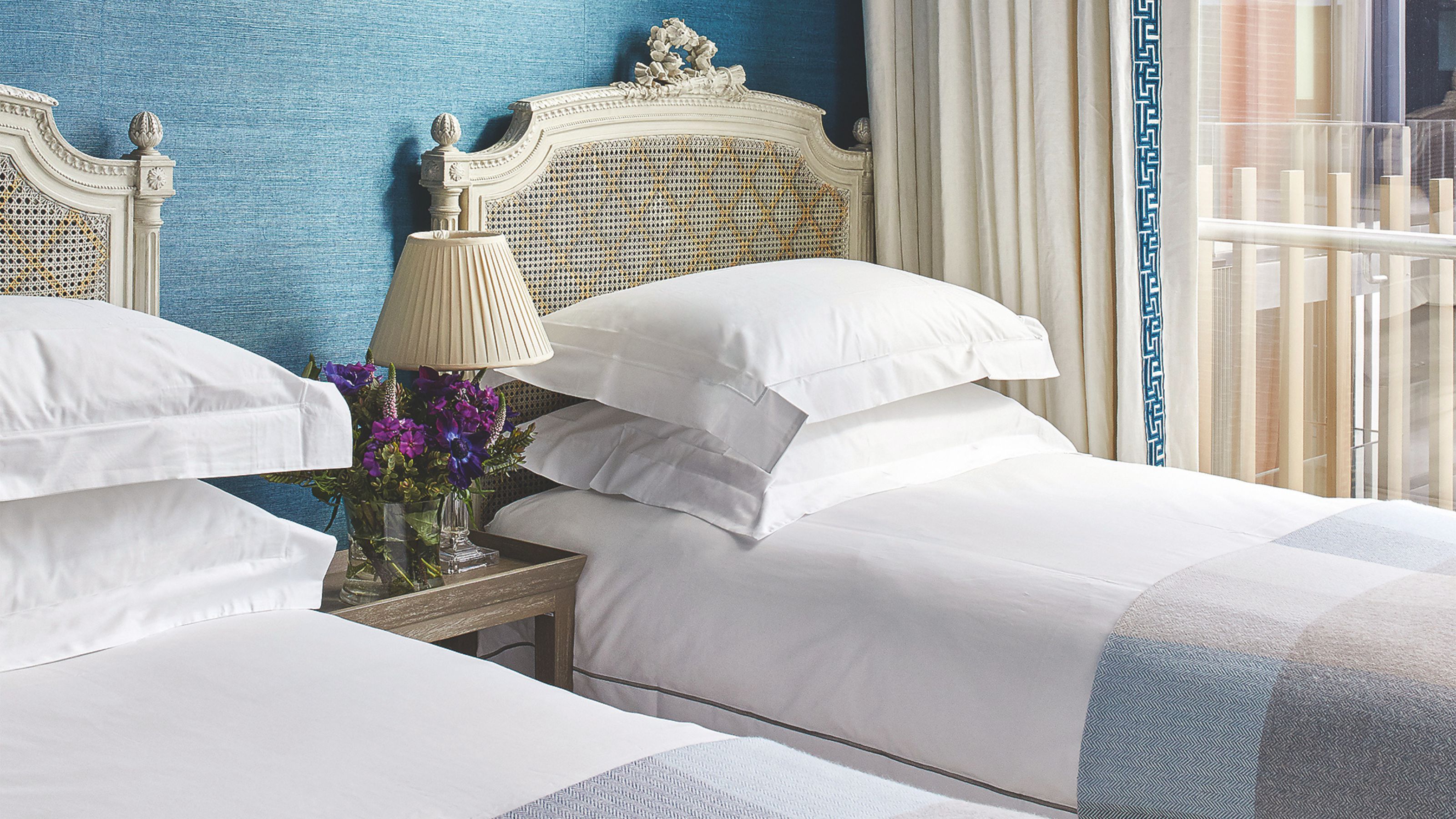 Blue twin bedroom with white rattan headboards and nightstand between with purple flowers and small lamp with white lampshade