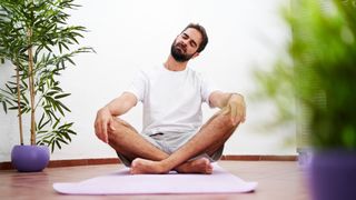 Man sits with crossed legs on a yoga mat and performs neck rotations. His hands are on his knees and his eyes are closed. He wears a white shirt, grey shorts and has dark hair and a beard.