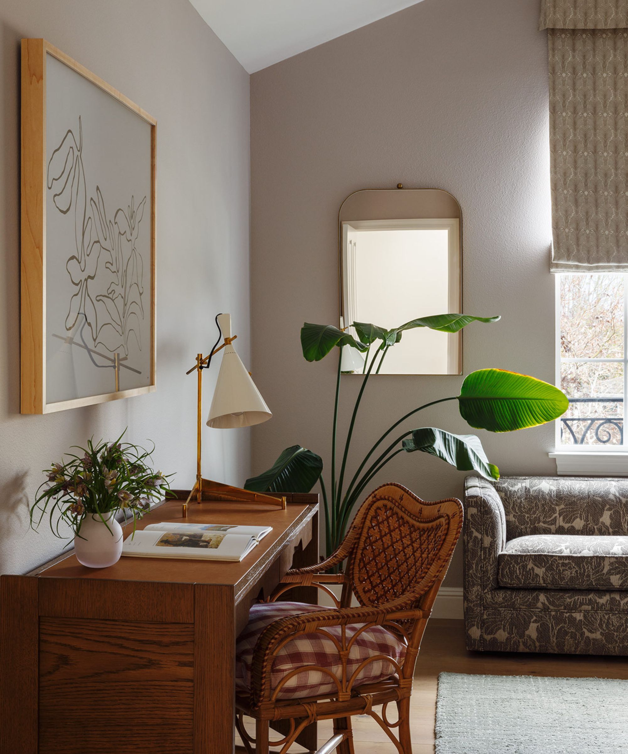 A bedroom with light mauve walls, a wooden desk with a vintage chair, a patterned sofa, a patterned roman blind, and a full-length mirror.