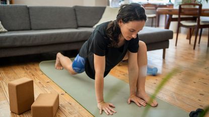 Woman stretches her hips in front of a couch with two yoga blocks to the side of her