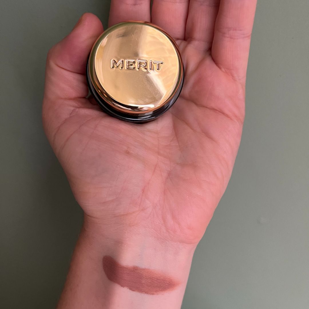 a hand on a green background holding Merit Solo Shadow cream eyeshadow with a swatch of the eyeshadow on the wrist 