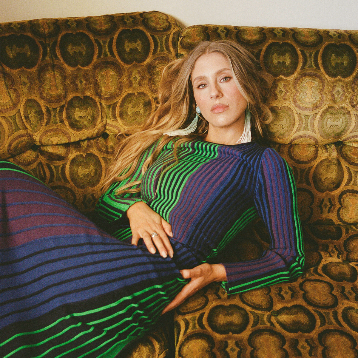 Lisa Gilroy photographed seated on a brown coach with circular pattern wearing a green, purple, and blue striped long sleeve dress and light blue fringe earrings. 