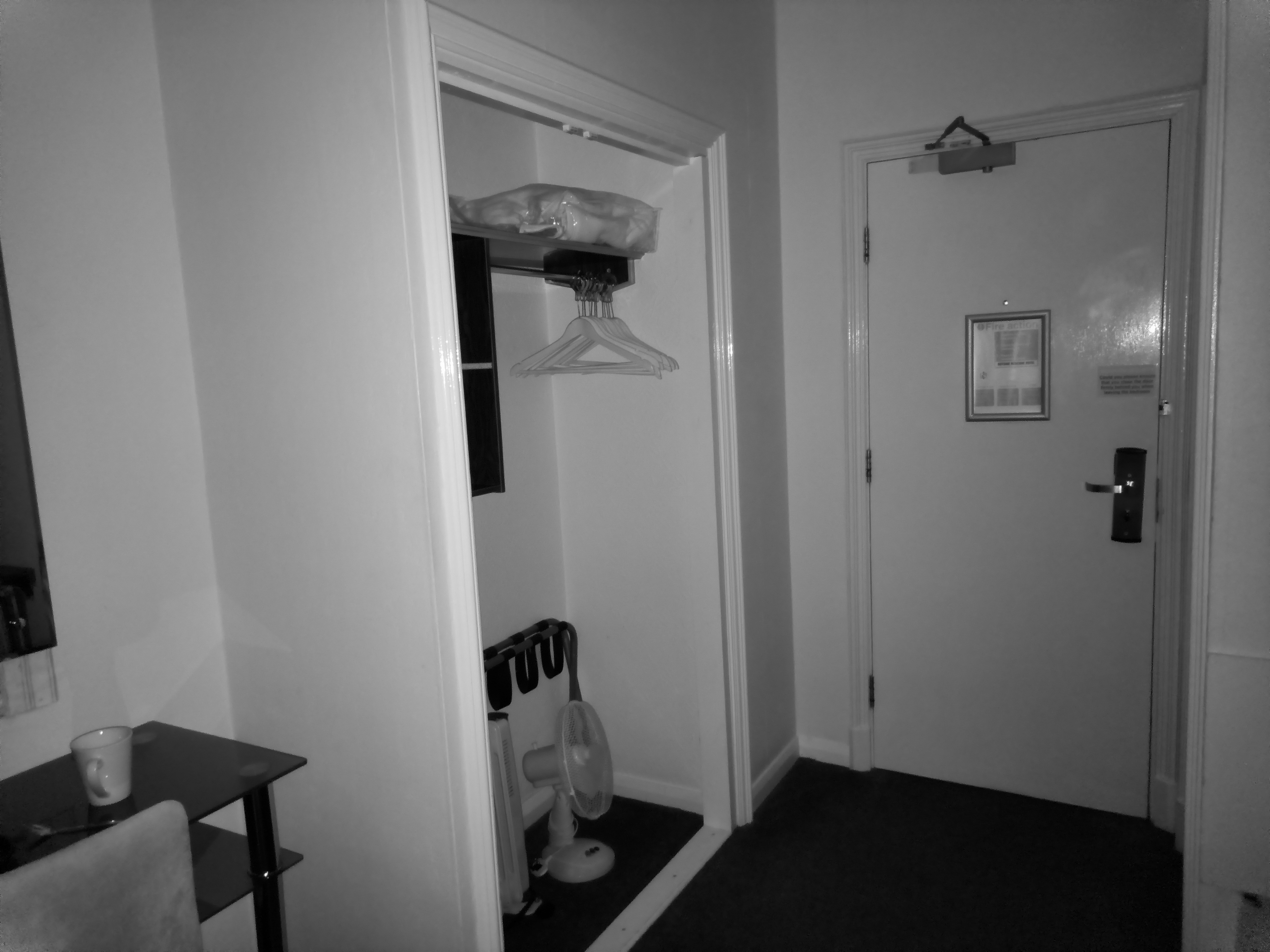 Black and white photo of a hotel room entryway showing an open closet with hangers, a small desk with a cup, and an entry door