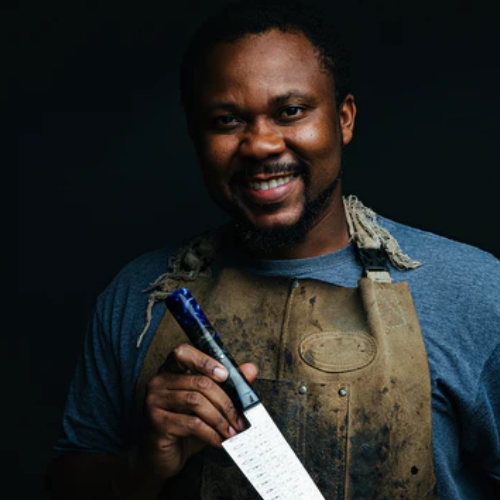 Image of a man in a blue t-shirt and apron holding a cooking knife.