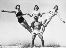 While Charles Atlas, who claims the title or the world's most perfect physique, is not carrying the entire globe on his shoulders might he be said to be carrying a fair part of it? Atlas was snapped during an early season workout on the beach supporting three women; they are, left to right, Doris Aschoff, Dolores Foley and Audrey Aschoff.