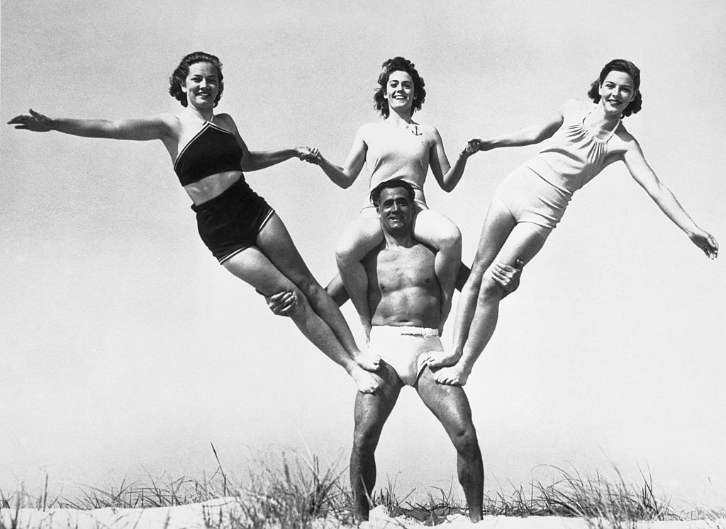 While Charles Atlas, who claims the title or the world's most perfect physique, is not carrying the entire globe on his shoulders might he be said to be carrying a fair part of it? Atlas was snapped during an early season workout on the beach supporting three women; they are, left to right, Doris Aschoff, Dolores Foley and Audrey Aschoff.
