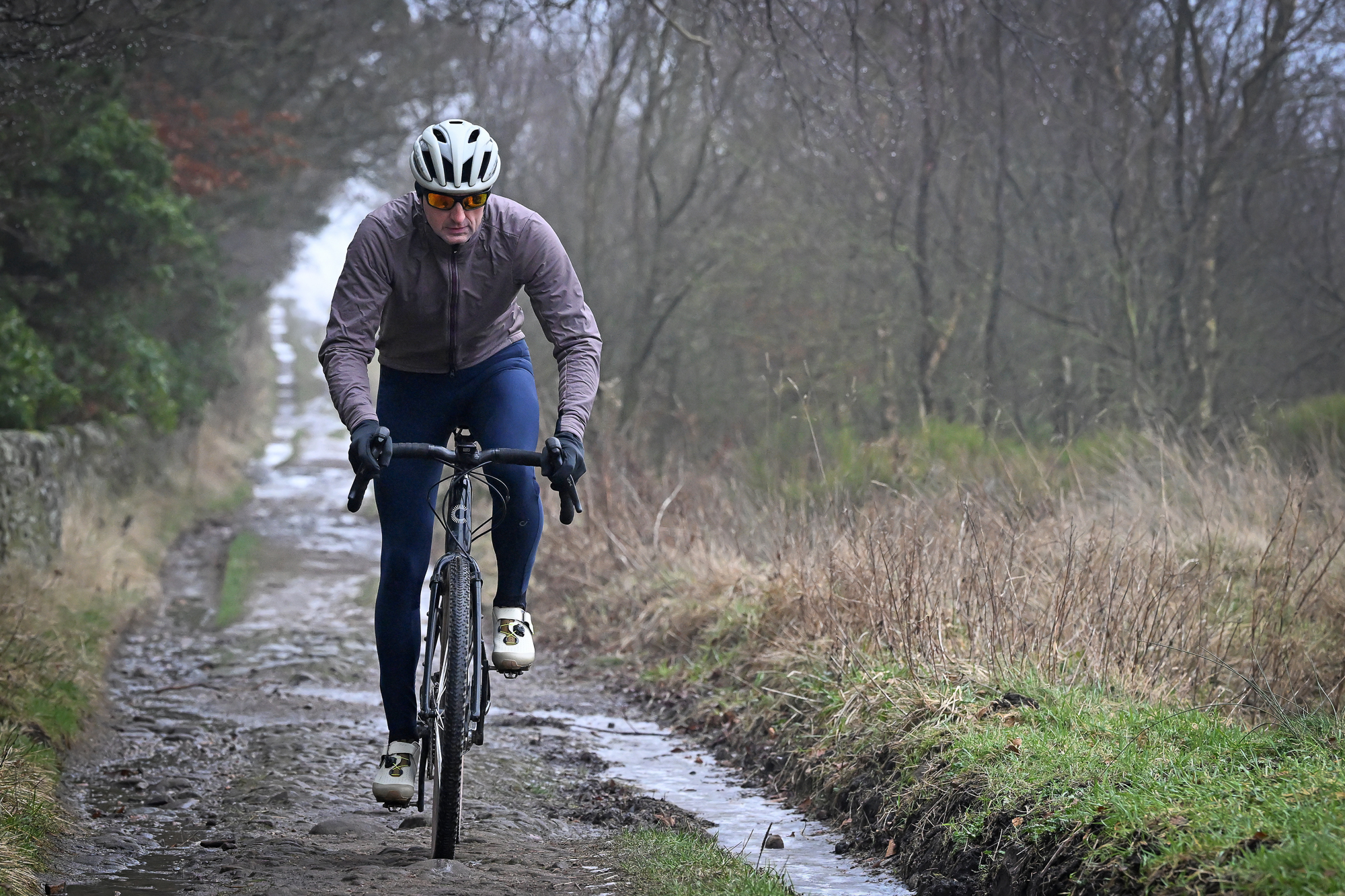 Man riding a gravel bike towards the camera on a dirt track wearing blue tights, purple jacket and beige shoes
