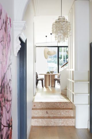 White hallway with peach terrazzo tile stairs leading into a modern dining room. There is a cream shell chandelier and a dark blue door frame in the hallway.