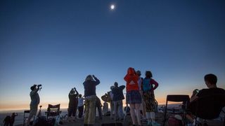 a crowd of people in a field look up at a solar eclipse taking place in a clear sky above