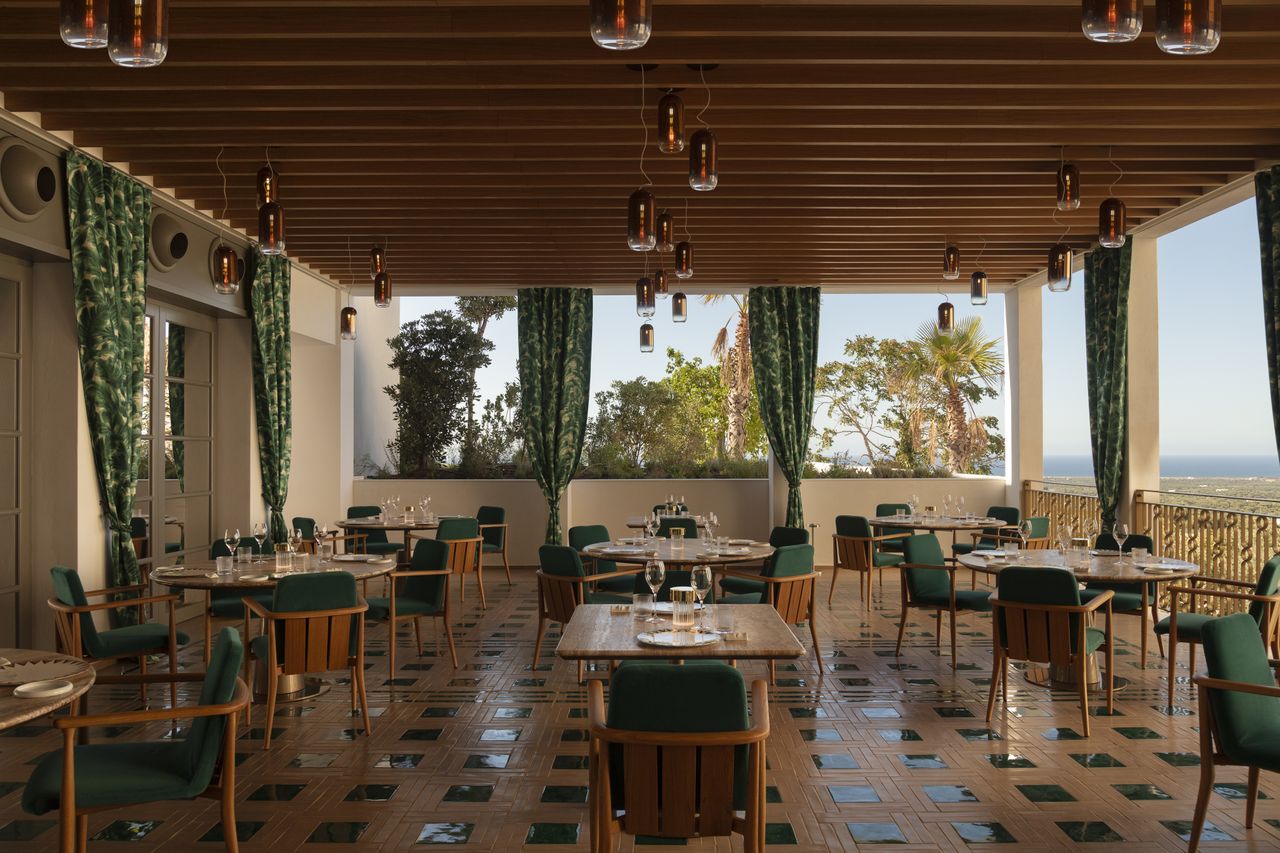A sea-view restaurant with checkered green and terracotta floors, emerald green and wood decor, and flowy green curtains.