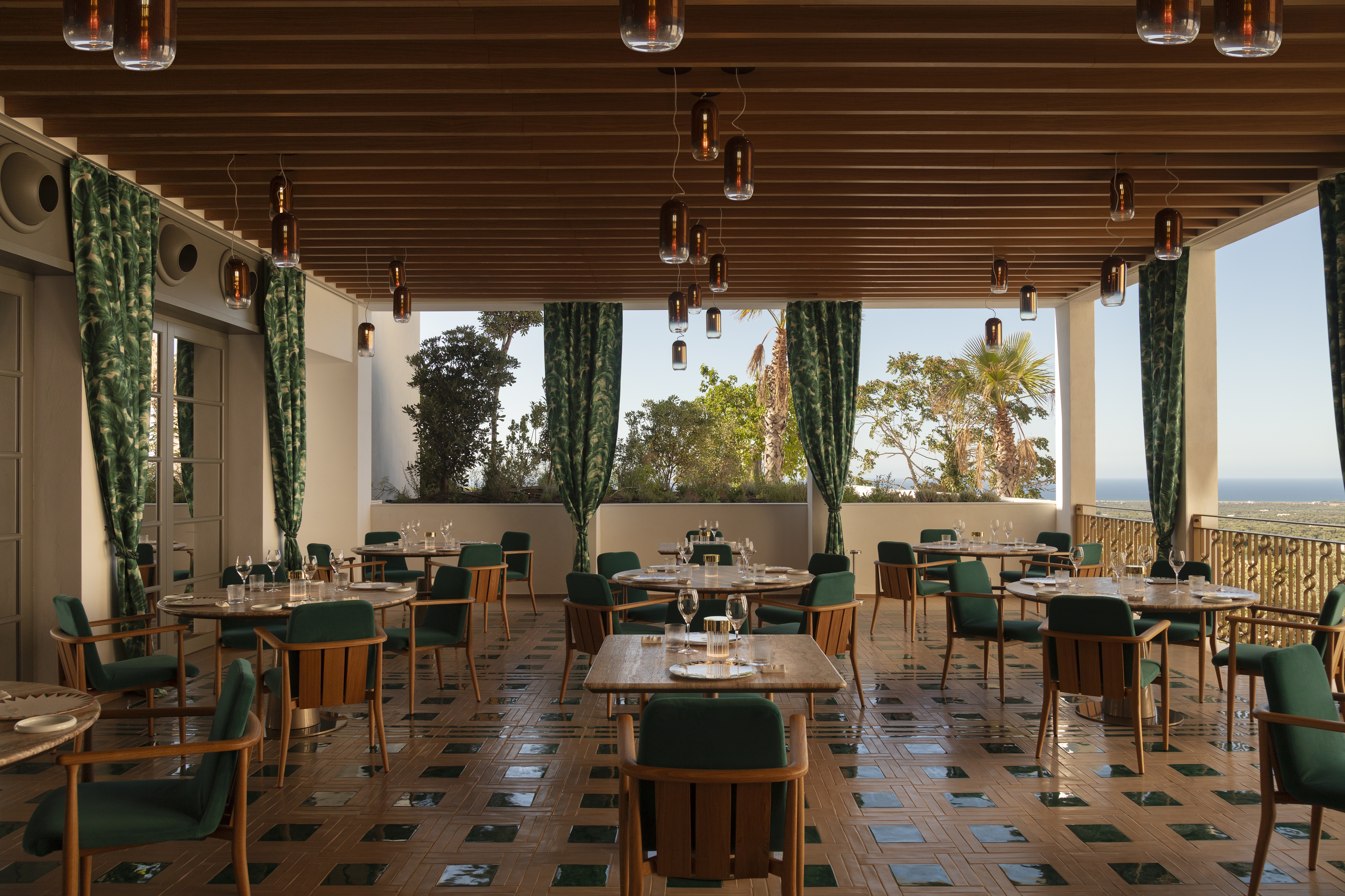 A sea-view restaurant with checkered green and terracotta floors, emerald green and wood decor, and flowy green curtains.
