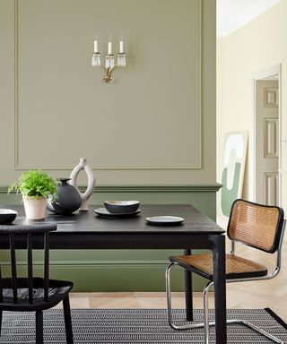 Green dining room painted two shades from little greene, wall paneling on walls, black wooden dining table, cesca chair in black and rattan, dining table dressed with ornaments, plates and plants, candelabra on wall