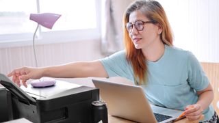 Woman using laptop reaching out towards a printer.