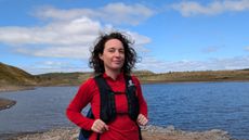 A woman hiker in front of a lake wearing a red fleece and a black hydration vest