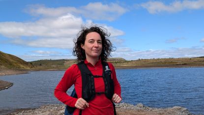 A woman hiker in front of a lake wearing a red fleece and a black hydration vest