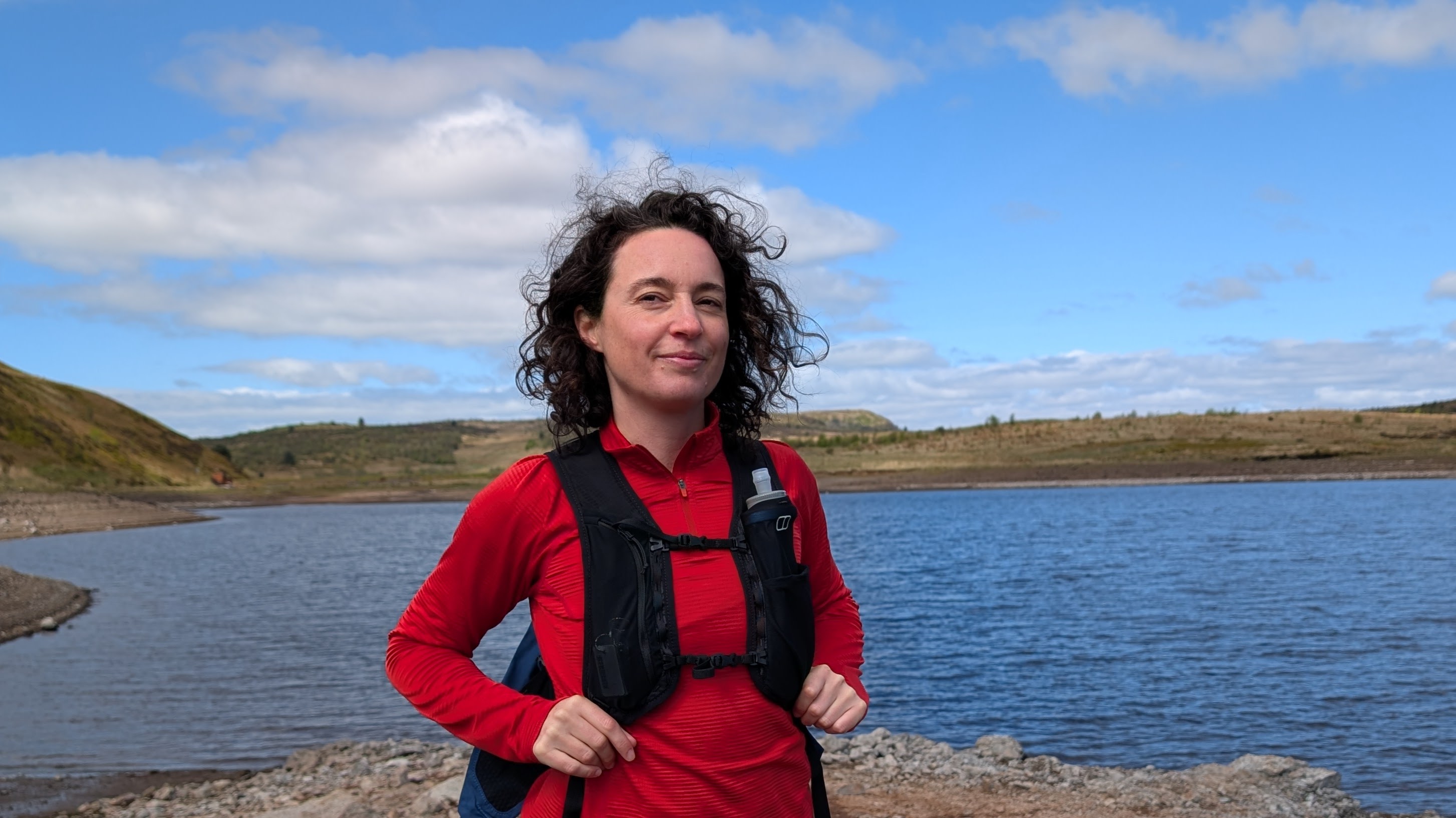 A woman hiker in front of a lake wearing a red fleece and a black hydration vest