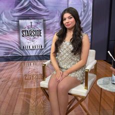 Alex Aster doing press for her new book Starside. She is sitting in a chair wearing a sequin floral embellished dress. 