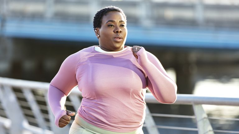 What does running do to your body: Pictured here, a woman in her 30s exercising in a city park on the waterfront. She is a plus size model with a large build, wearing long sleeved sports clothing. She is running, jogging or power walking toward the camera.
