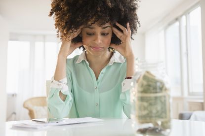 Woman sitting with head in her hands next to a jar containing money notes