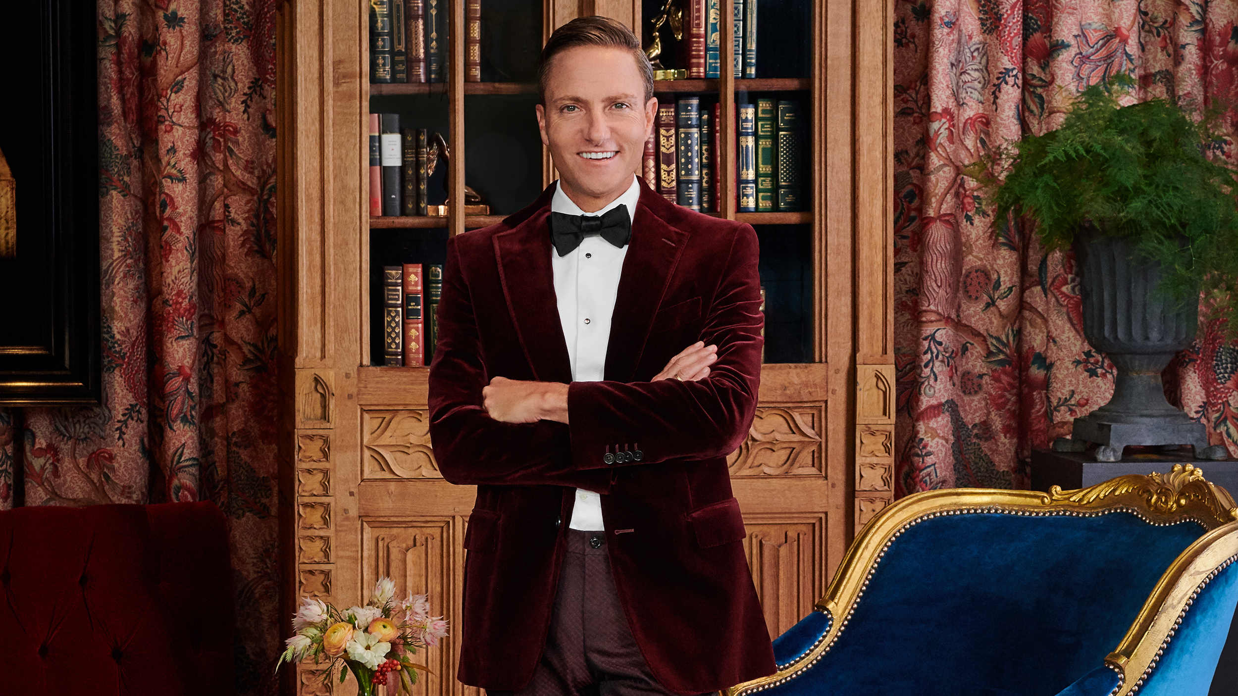 Ken Fulk in a red smoking jacket standing in front of an opulent curtain and bookcase