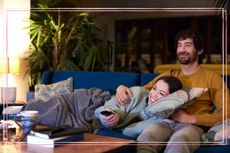 A couple cuddling on a sofa and pointing a remote at a TV