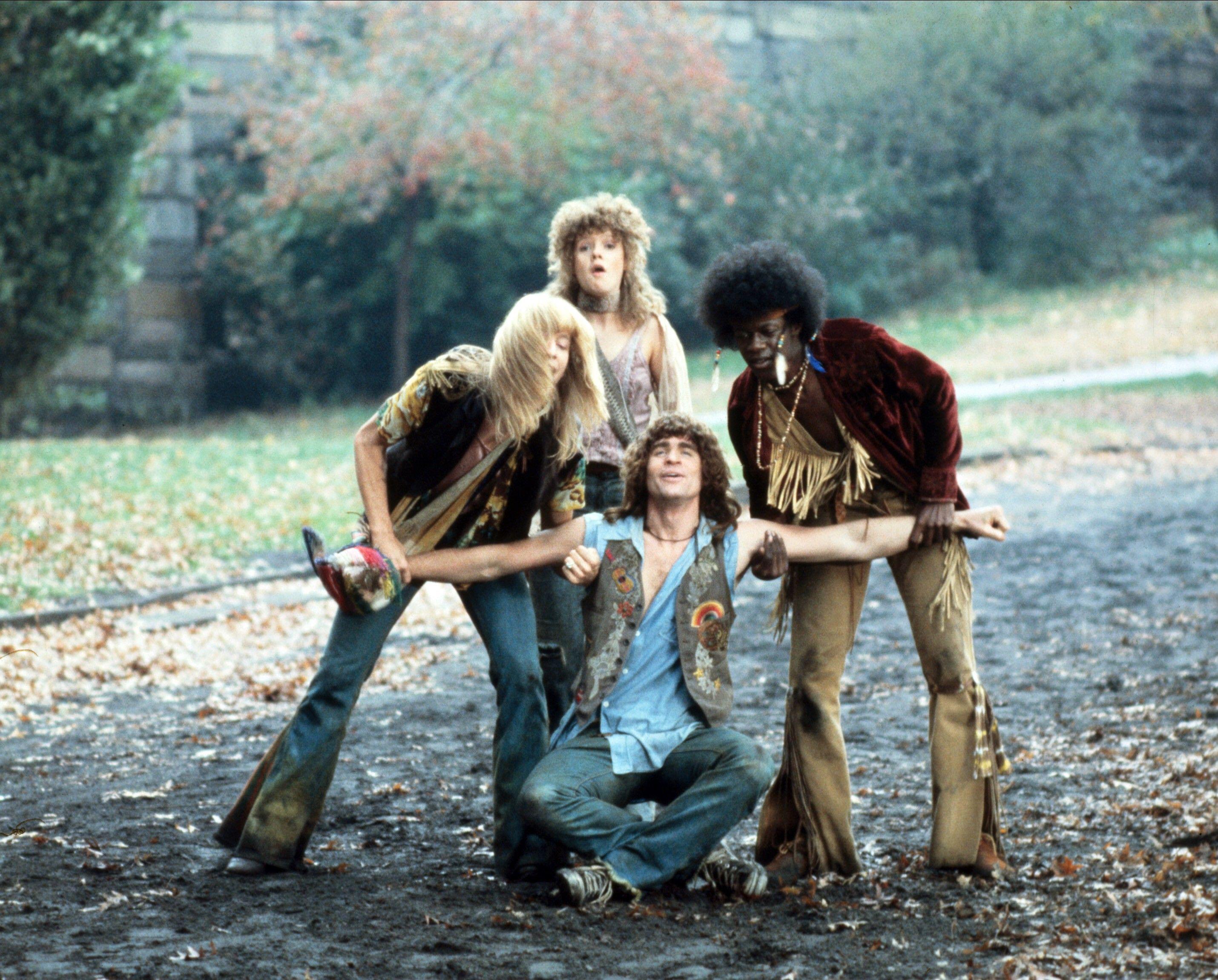 a group of 70s era hippies stand together while one sits on the ground with his arms stretch out in a still from the movie hair