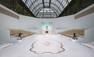 Catwalk viewing auditorium, white marble floor with gold motif, row of wooden tiered stools, white walls, two black cameras on tripods, gold framed catwalk entrance, metal framed arch ceiling with windows