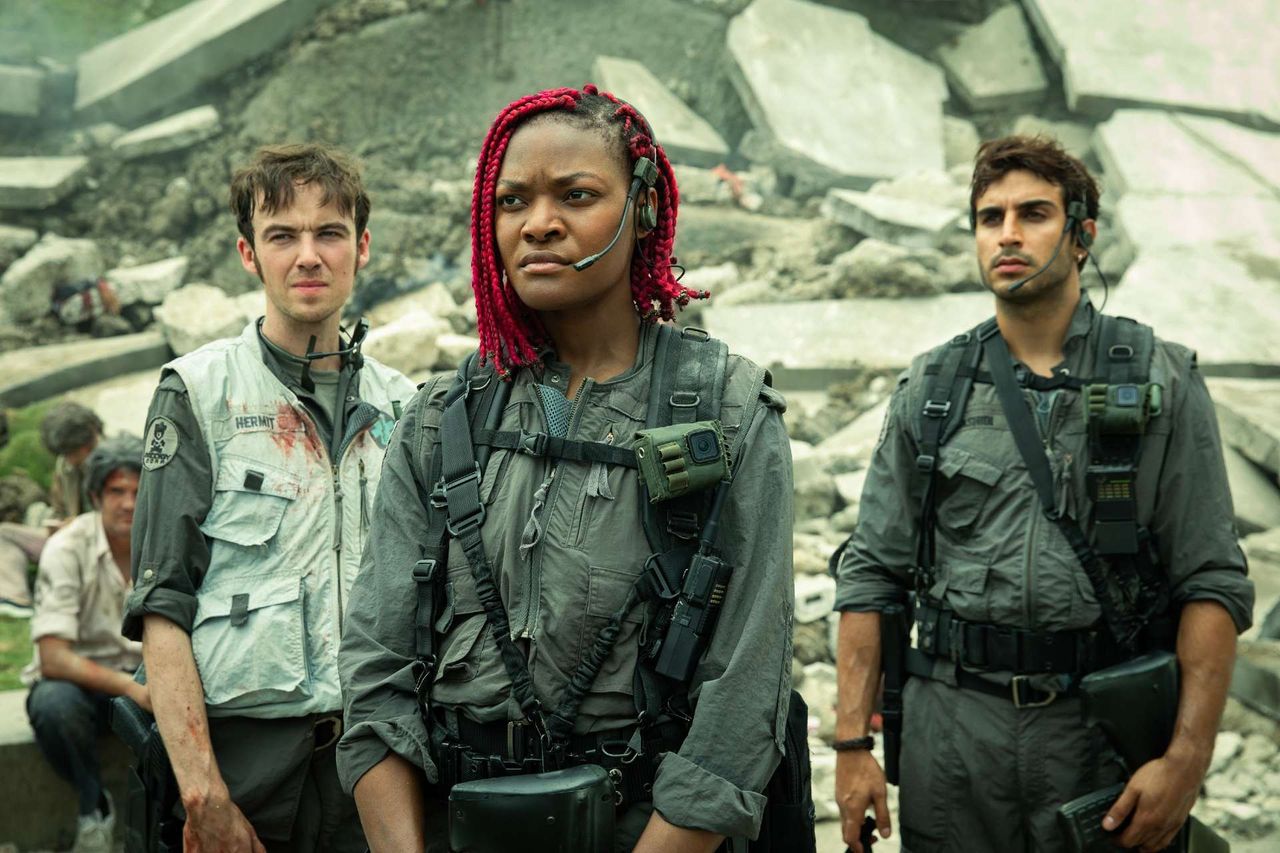Still from the TV show Alien: Earth. Three people are standing together in front of a large pile of rubble. They are each wearing a gray jumpsuit, utility belt, backpack, and a headset with a mic.