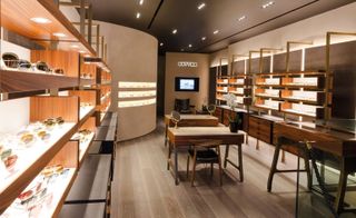 An eye glasses store with wooden shelves displaying many pairs of glasses and wooden tables with round mirrors on them in the middle of the room.