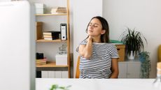 Woman holds her neck with her eyes closed. She is sitting at a desk in front of a computer screen.