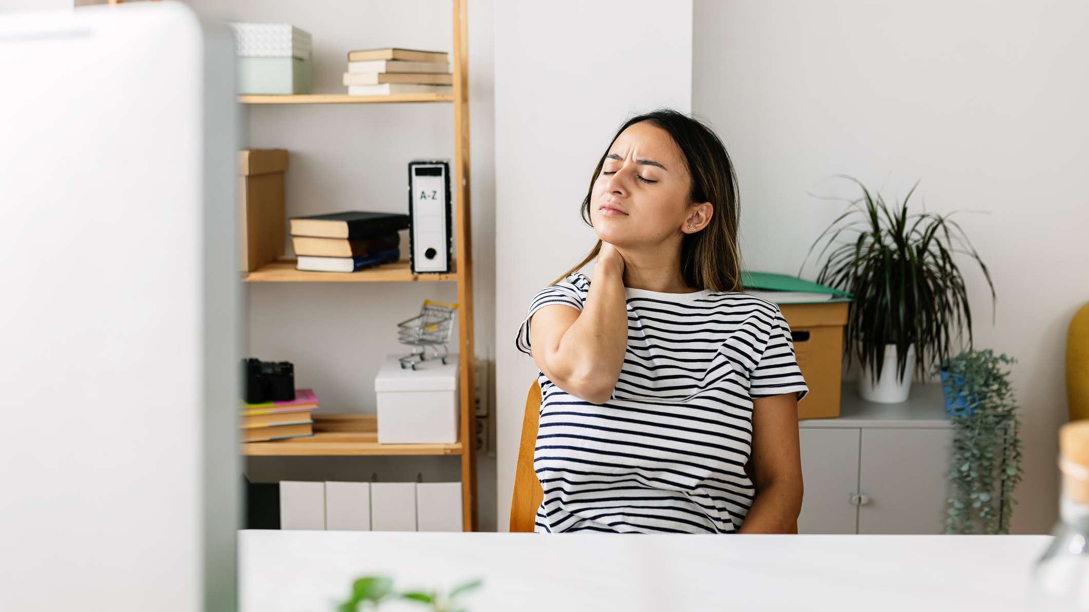 Woman holds her neck with her eyes closed. She is sitting at a desk in front of a computer screen.