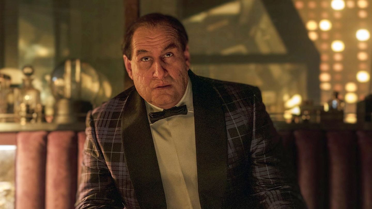 Scene from The Penguin. A man with slicked-back brown hair and a scarred face wears a black and purple plaid suit jacket with a black bow tie, looking directly at the viewer with a serious expression.