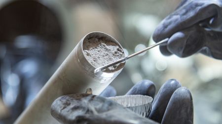 a close-up of two gloved hands holding a tube full of grey rocky dust in a clean room