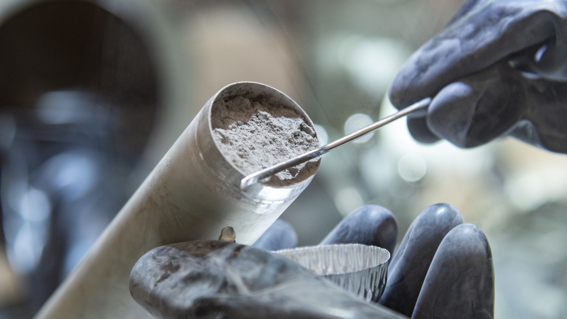 a close-up of two gloved hands holding a tube full of grey rocky dust in a clean room