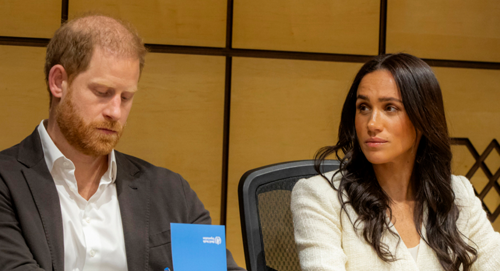 Prince Harry wears a suit without a tie and looks somber while wife Meghan Markle wears a white top and has long black hair