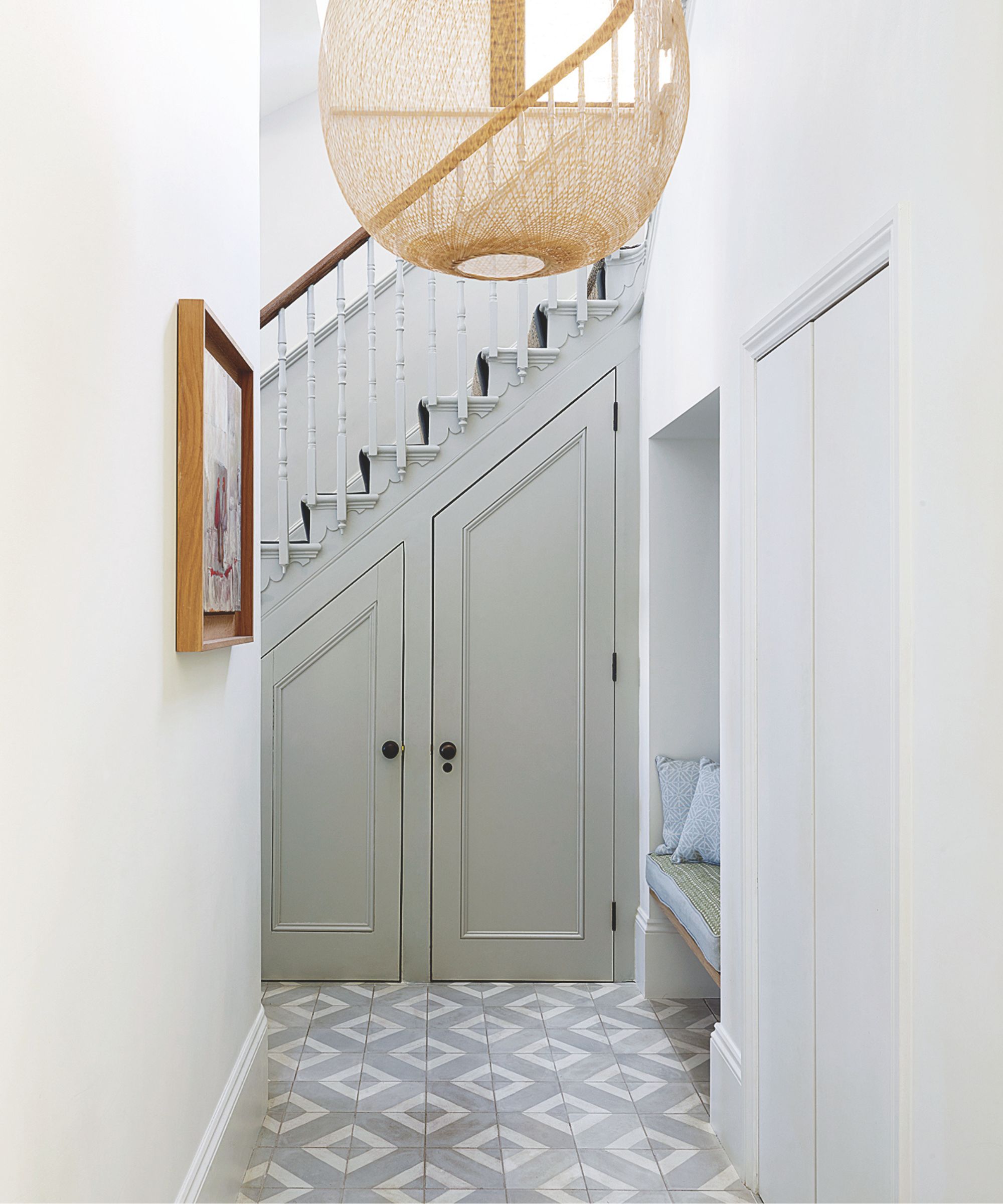grey painted sloped staircase with traditional spindles and shaker cupboards underneath with a large woven wicker ceiling light and geometric floor tiles
