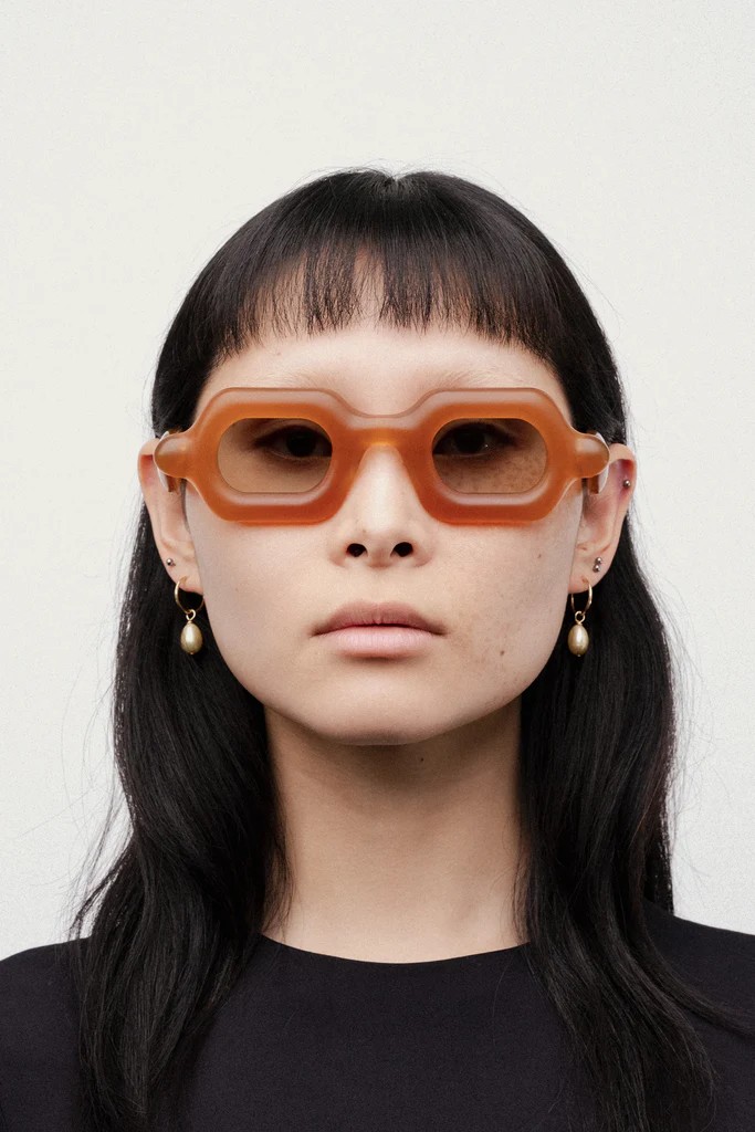 A woman with black straight hair and a fringe wears a pair of golden pendant earrings and toffee-hued, chubby-looking sunglasses.