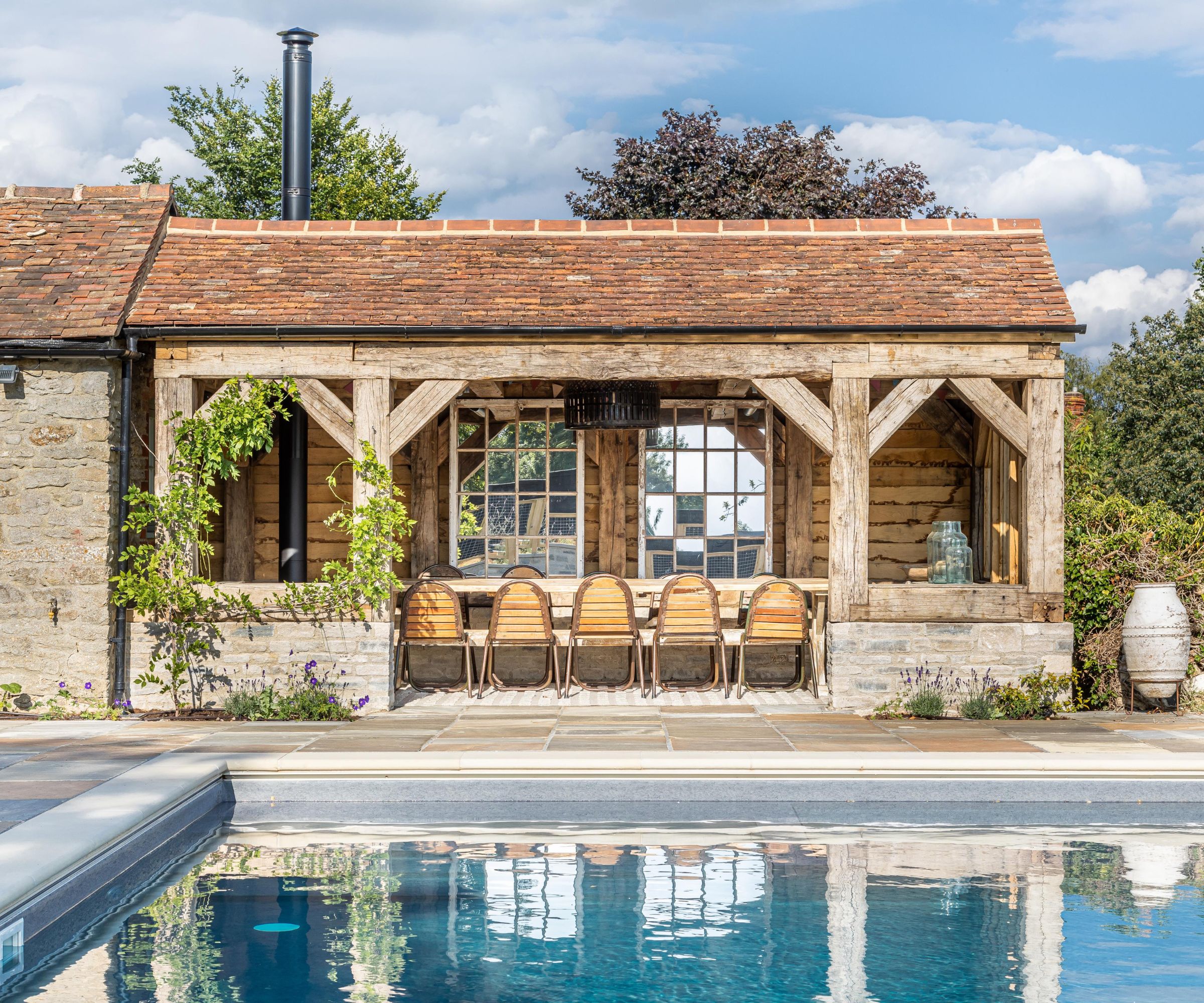 A rustic open-sided oak frame pool house with a clay tile roof, antique glazing and a long teak dining table, beside a slate-edged pool with stone flags and lavender planting.