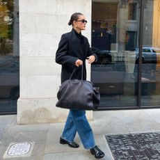 Influencer @sobalera stands outside wearing Oxford shoes, blue jeans, an oversized blazer and a large black handbag.