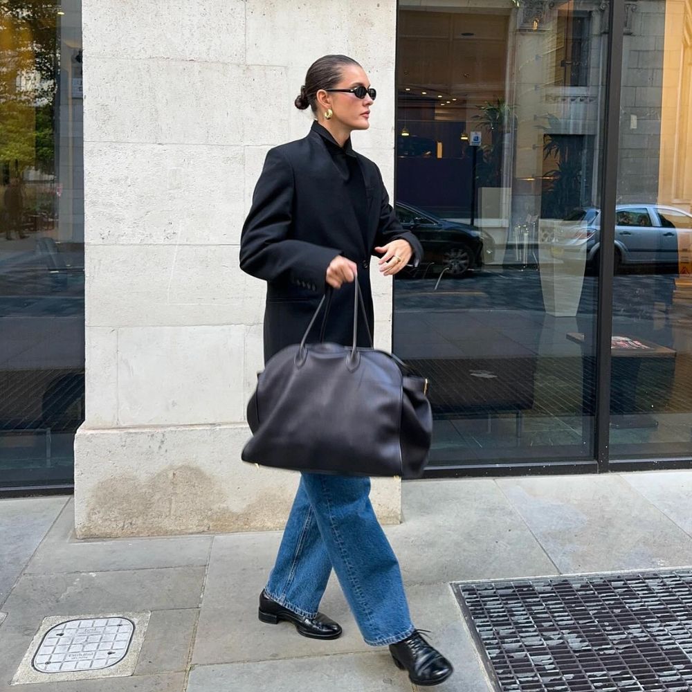 Influencer @sobalera stands outside wearing Oxford shoes, blue jeans, an oversized blazer and a large black handbag.