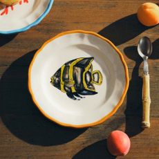 A fish, colourful, embroidered plate with a colourful table setting with a spoon and peaches from Mytheresa Life