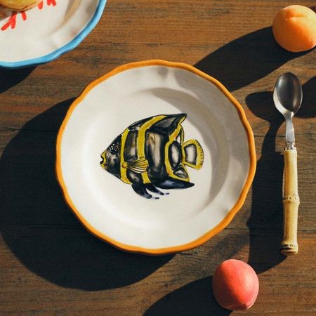 A fish, colourful, embroidered plate with a colourful table setting with a spoon and peaches from Mytheresa Life