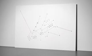 A large white rectangular board placed against a white wall with threads moving around pins in a geometric pattern