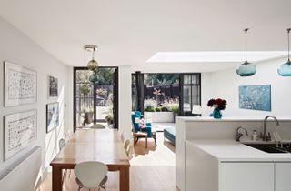 large white kitchen extension with contemporary finish by mulroy architects, including a long wooden dining table, two doors with black trim out to the garden, and white countertops and cabinets, and a blue armchair beyond