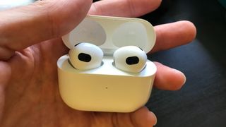 En hand håller upp ett par Apple AirPods 3 som ligger i sitt tillhörande laddningsfodral.